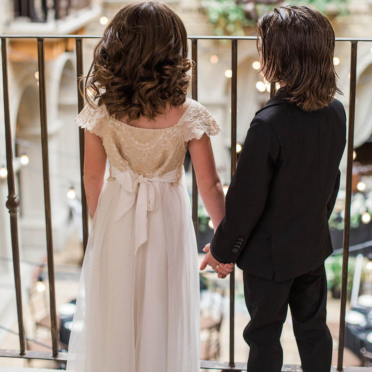 Deux enfants en tenue de cérémonie, main dans la main, regardant vers la salle décorée.