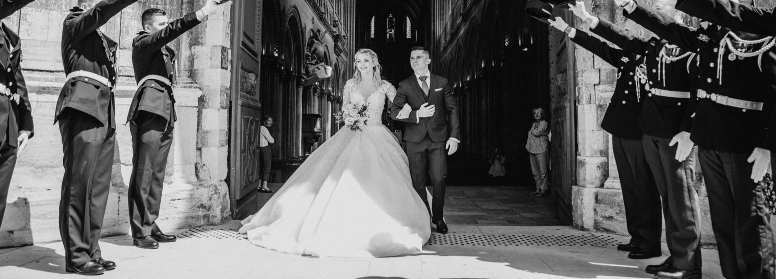 Mariée en robe princesse sortant de l’église aux côtés du marié, encadrés par une haie d’honneur militaire, photo en noir et blanc 